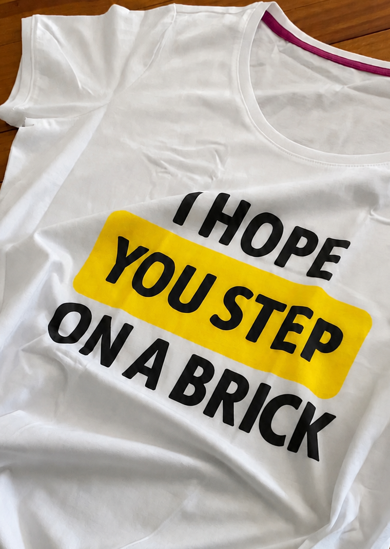White T-shirt with the words “I hope you step on a brick” lying on a wooden table. The words “You step” are highlighted in yellow.